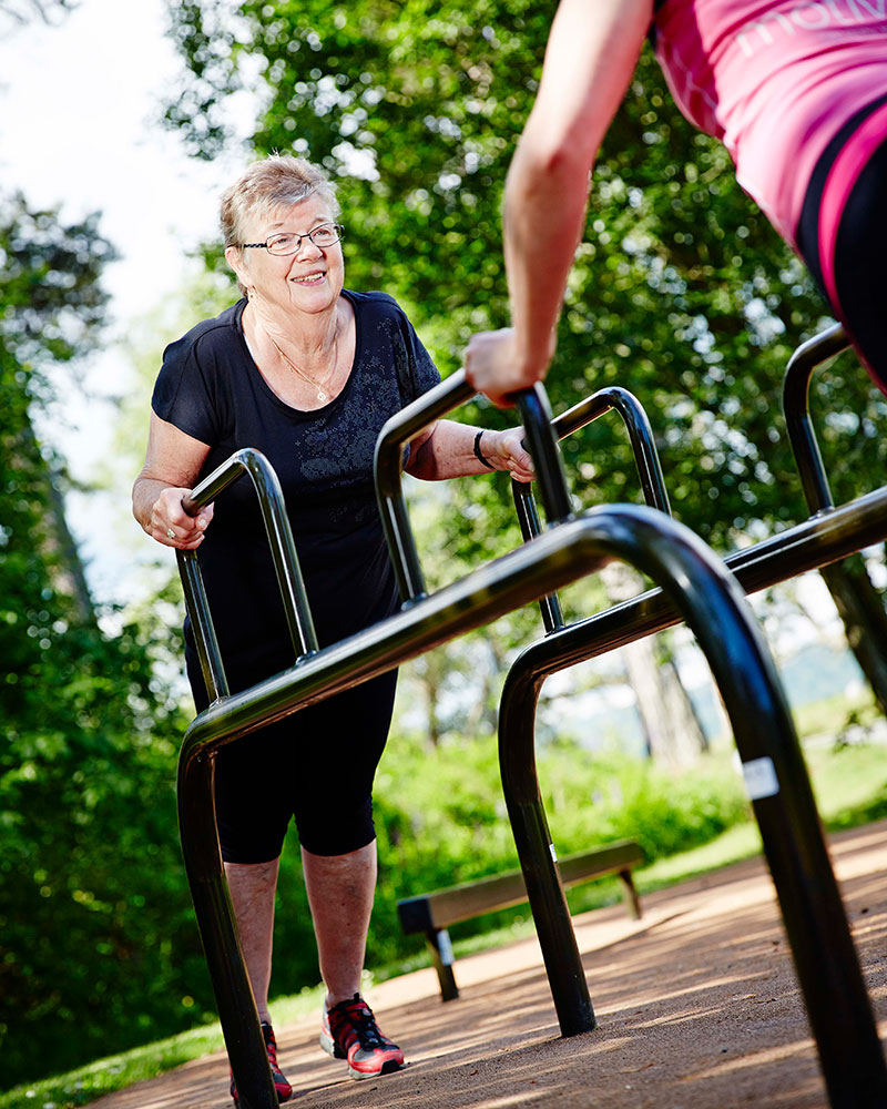 An elderly woman completes dips at an outdoor fitness area, she is looking up and smiling.